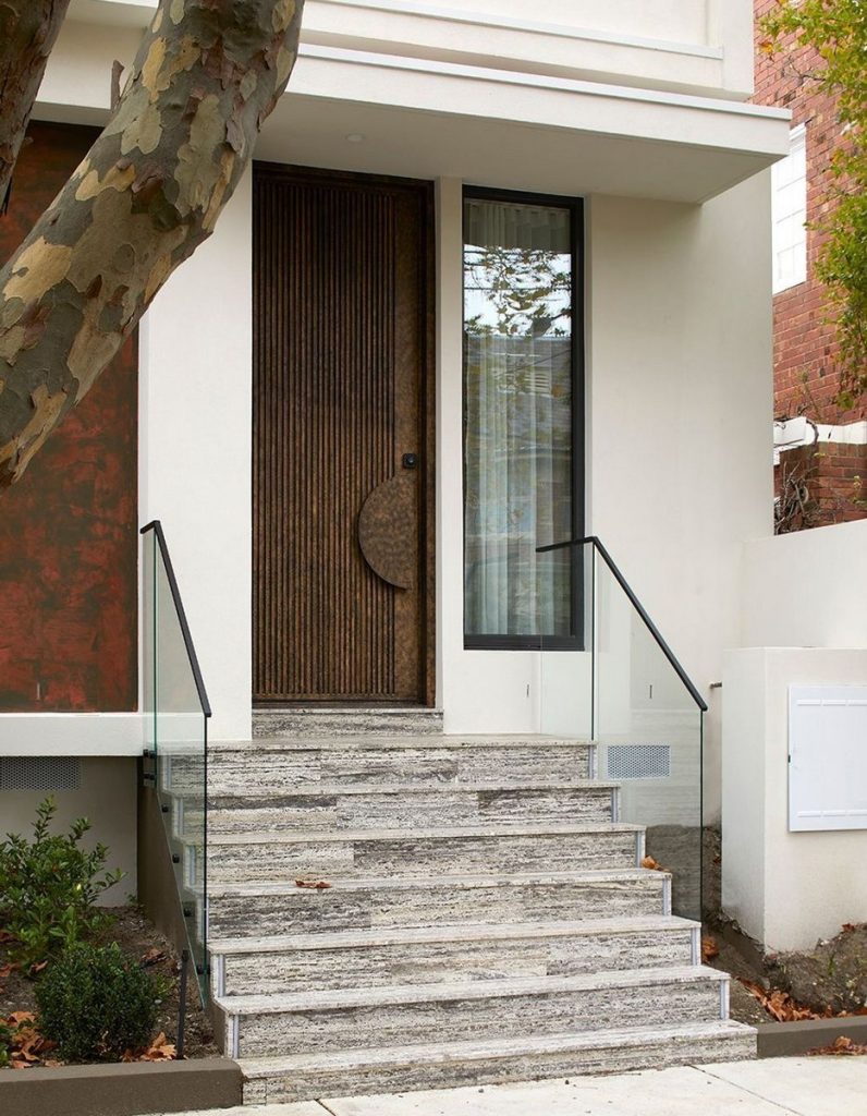 Modern entrance featuring a textured wooden door, large glass window, grey stone steps, and sleek glass railings, framed by foliage.