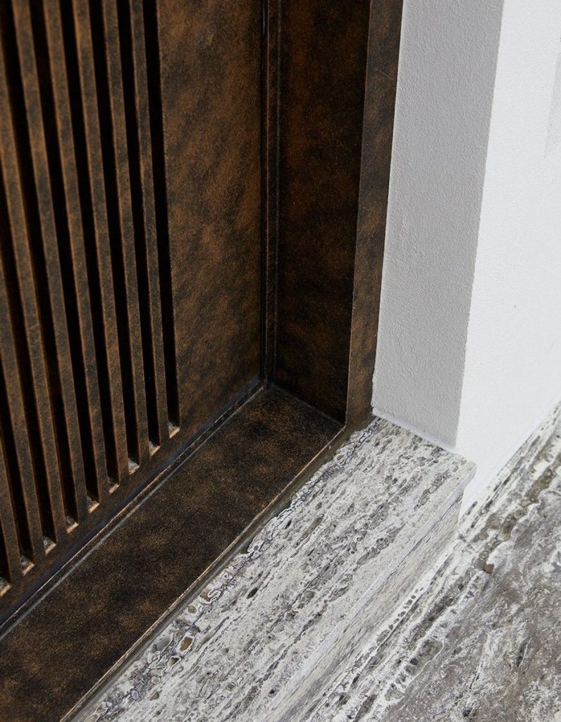 Close-up of a bronze-finished steel door with vertical slats, set against a textured stone base and white wall, showcasing elegant design.