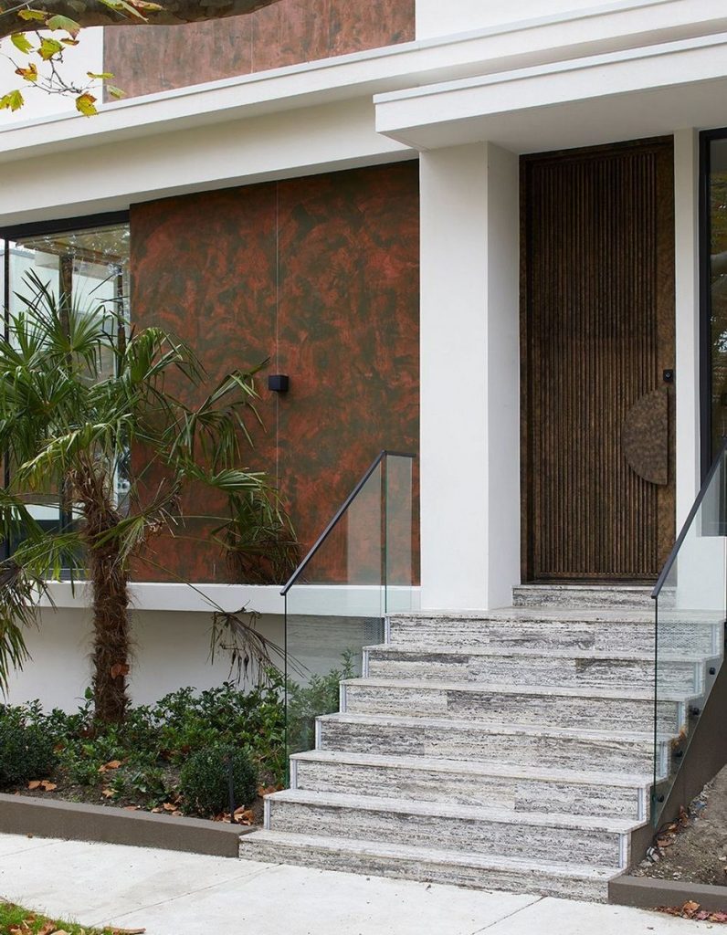 Contemporary entrance featuring textured wood door, glass handrail, and stone steps, surrounded by greenery and a rust-coloured wall.