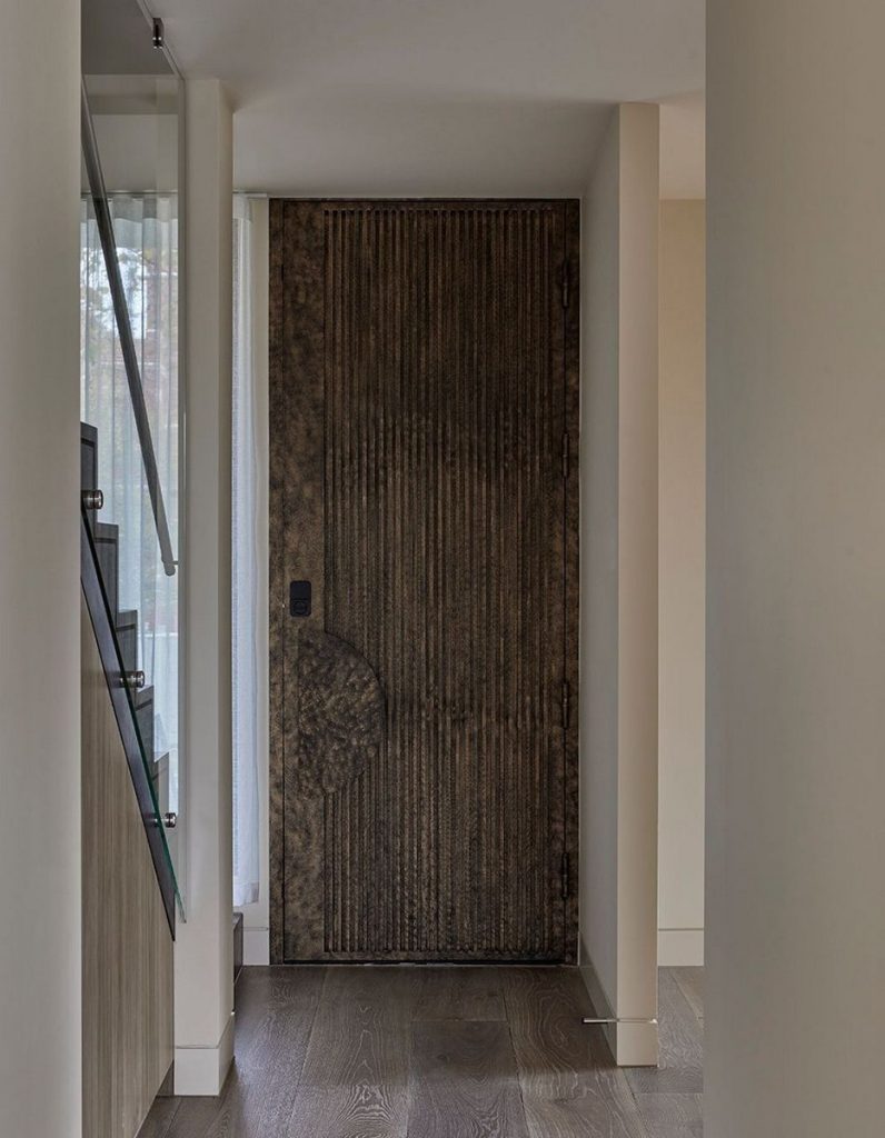 Bespoke steel door with a textured, dark finish, framed by minimalist interior walls and a glass staircase, enhancing modern elegance.