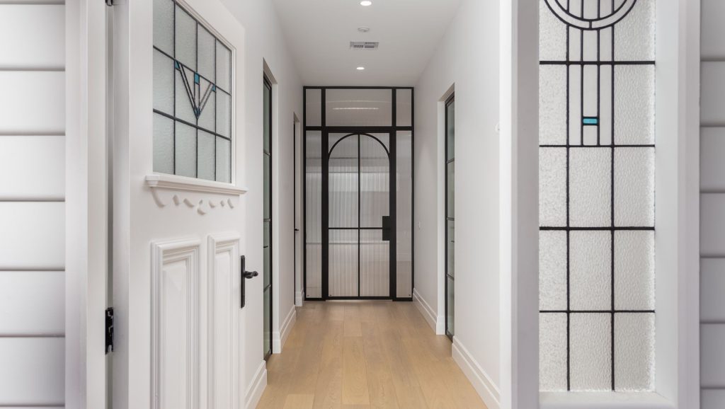 Hallway featuring a white door with ornate detailing, flanked by glass panels, leading to a black steel-framed archway and bright interior.