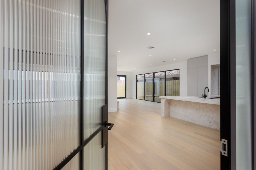 Steel-framed glass doors open to a spacious interior with light wood flooring, a stone island, and expansive glass walls leading outside.