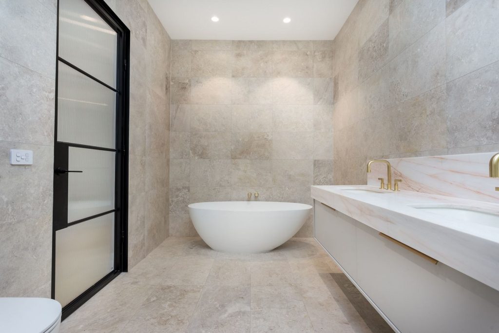 Spacious bathroom featuring a freestanding white bathtub, dual sinks with gold fixtures, and a glass-panelled black door against stone tiles.
