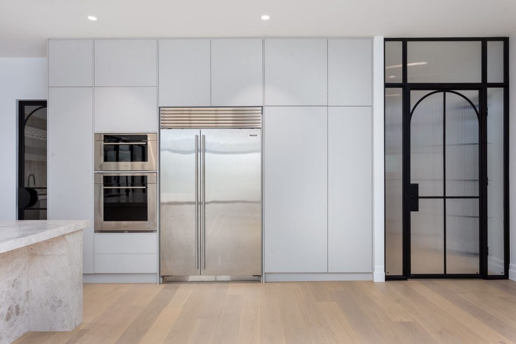 Modern kitchen featuring integrated white cabinetry, stainless steel refrigerator, built-in oven, and sleek black-framed glass doors.