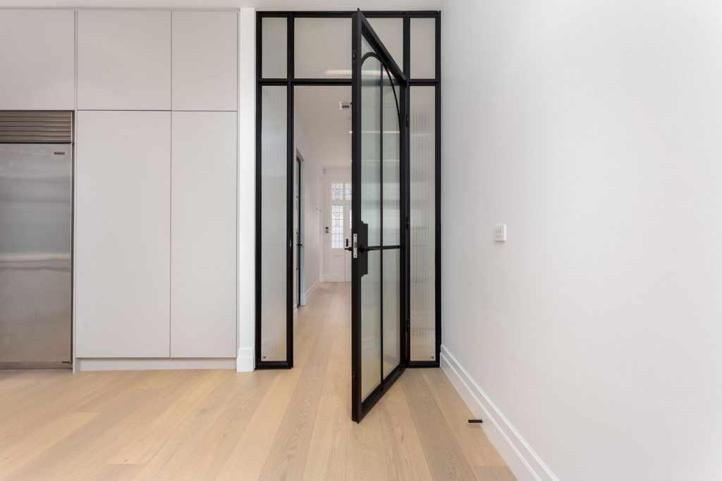 Modern interior showcasing a sleek black steel door, opening onto a light wooden floor, with minimalist white walls and cabinetry.