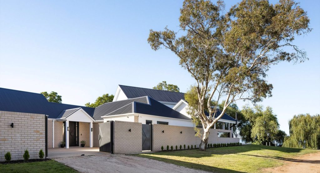 Modern residence featuring a mix of brick and steel accents, with landscaped grounds and a large tree, under a clear blue sky.
