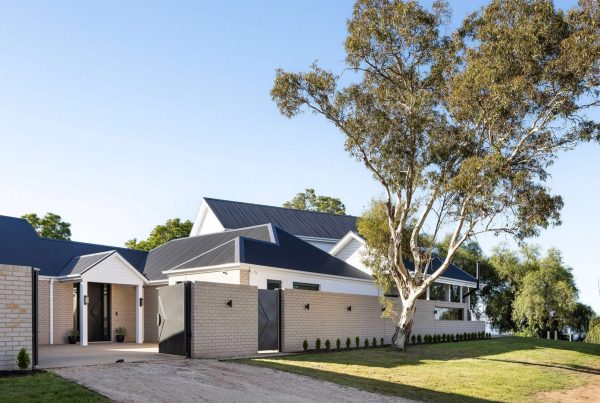 Modern residence featuring a mix of brick and steel accents, with landscaped grounds and a large tree, under a clear blue sky.