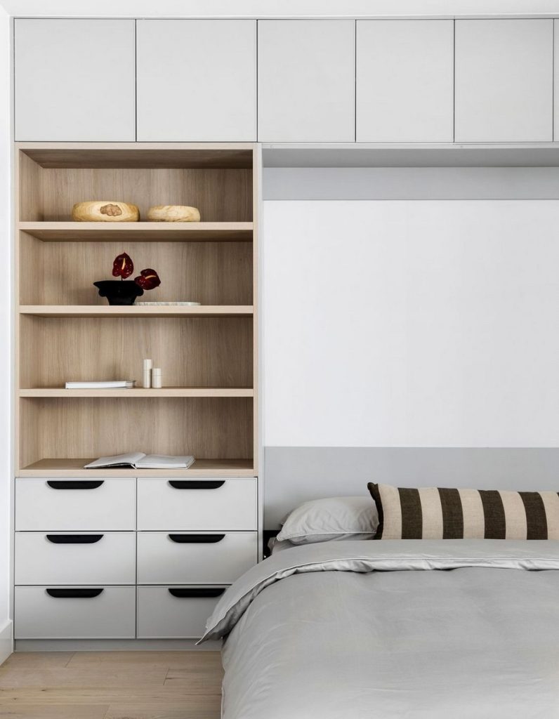 Modern bedroom featuring a light wood shelving unit with white cabinetry, displaying decorative items and a neatly made bed with striped cushions.