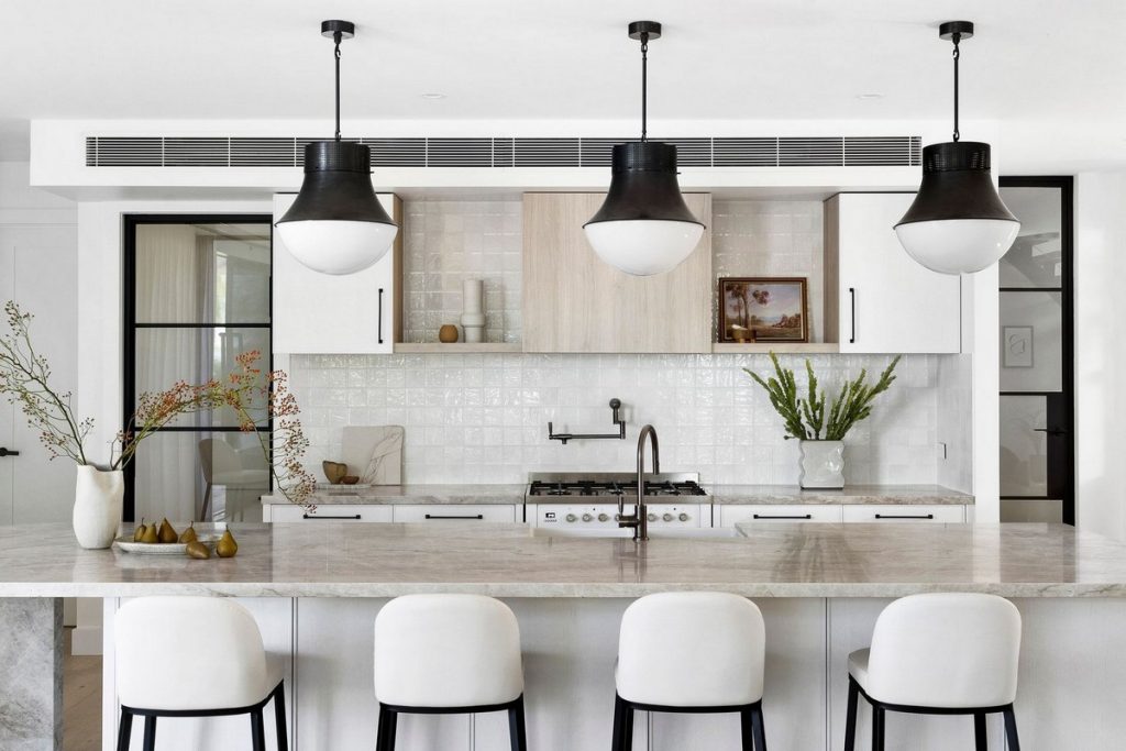 Modern kitchen with a marble island, white and wooden cabinetry, black pendant lights, and decorative plants, featuring neutral tones.