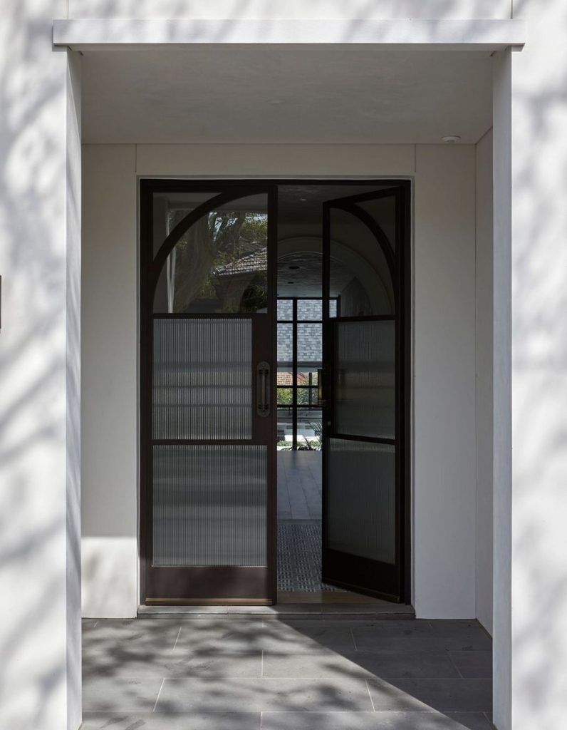 Bespoke steel-framed doors with textured glass panels open to a modern entryway, framed by minimalist architecture and natural light.