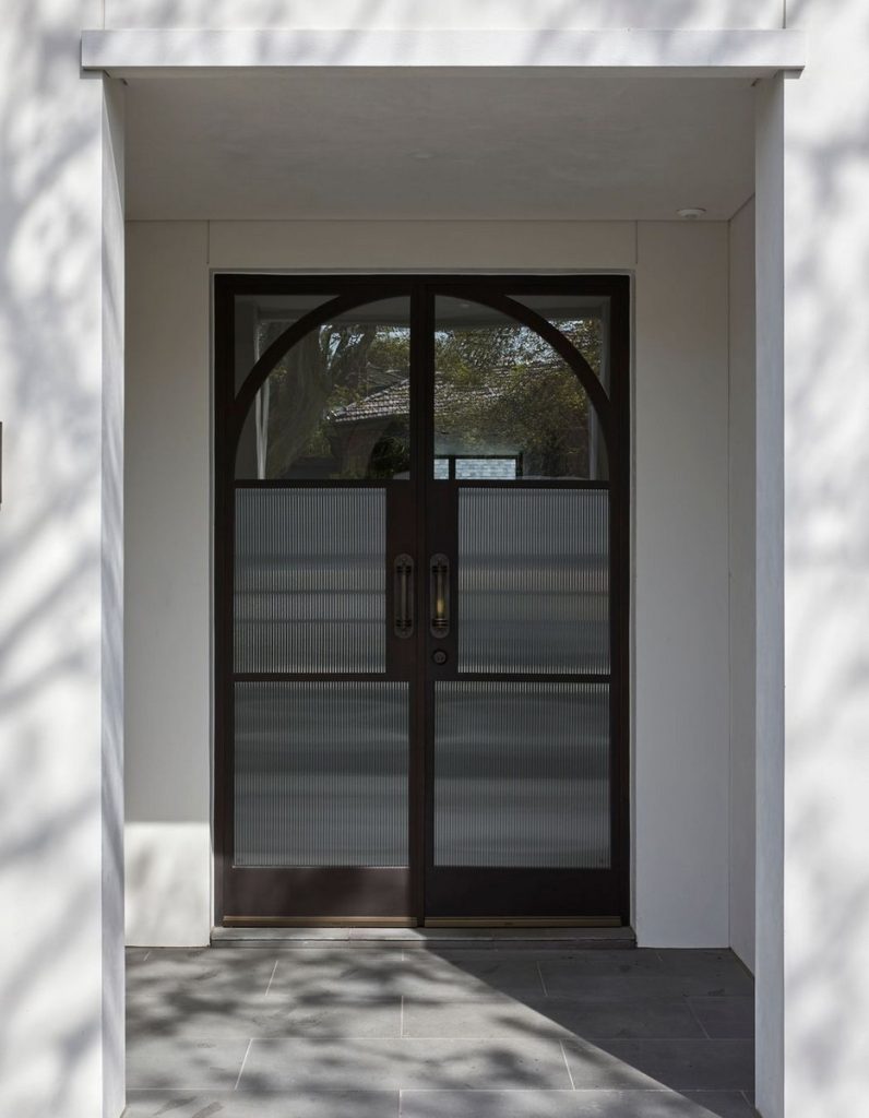 Bespoke steel door with an arched top and frosted glass panels, framed by a minimalist white entrance and shadowed by nearby trees.