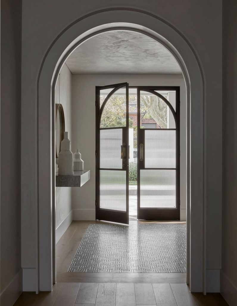 Elegant arched entrance showcasing bespoke steel doors with frosted glass, leading to a serene garden view with neutral tones.
