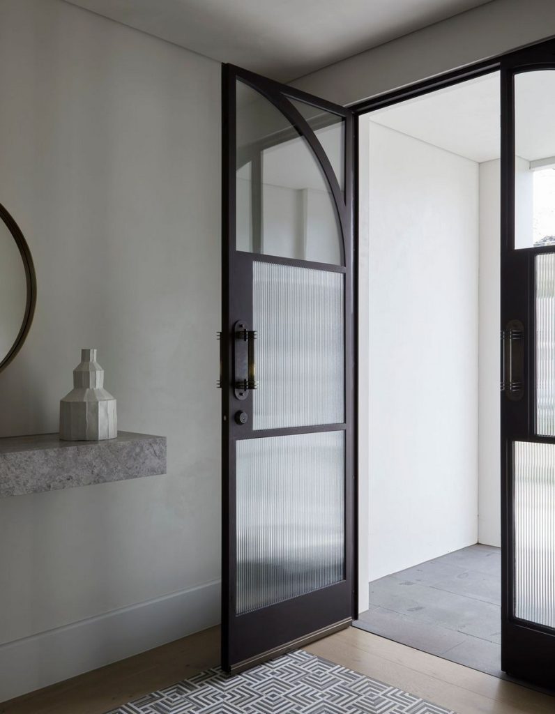 Bespoke steel-framed door opens into a minimalistic interior with light tones, showcasing a geometric patterned mat and a decorative vase.
