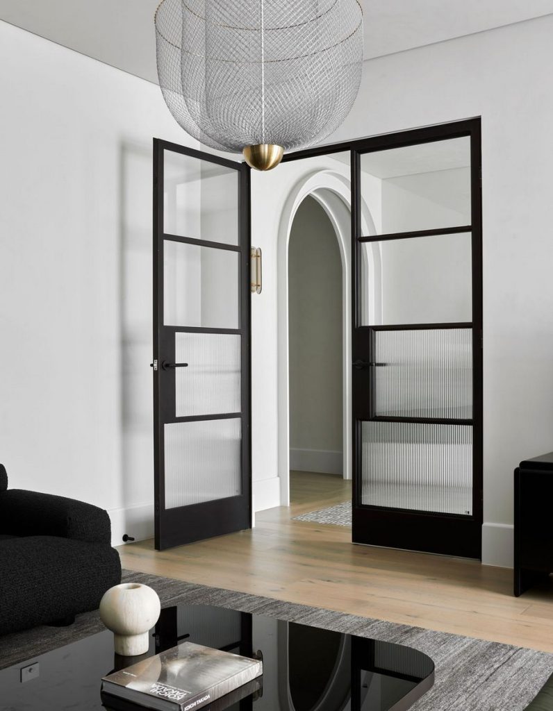 Black steel-framed doors open into a modern interior, featuring a textured glass design, neutral tones, and minimalist decor.