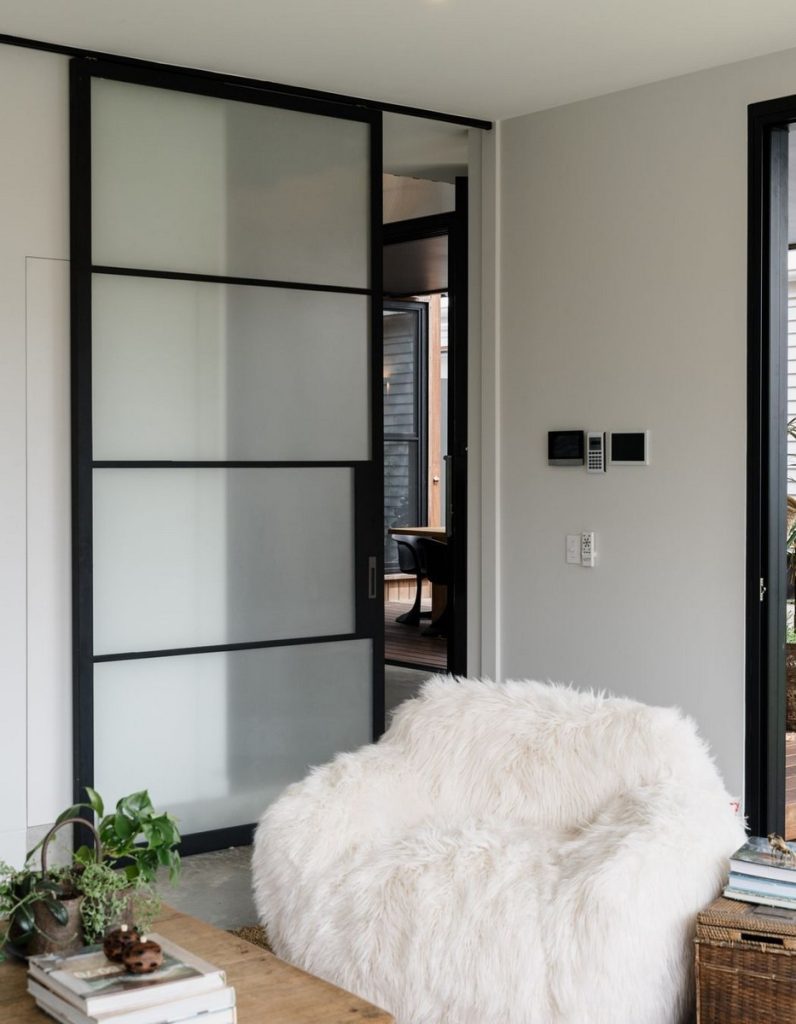 Bespoke steel sliding door with frosted glass panels divides a contemporary living space, featuring a textured white armchair and natural elements.