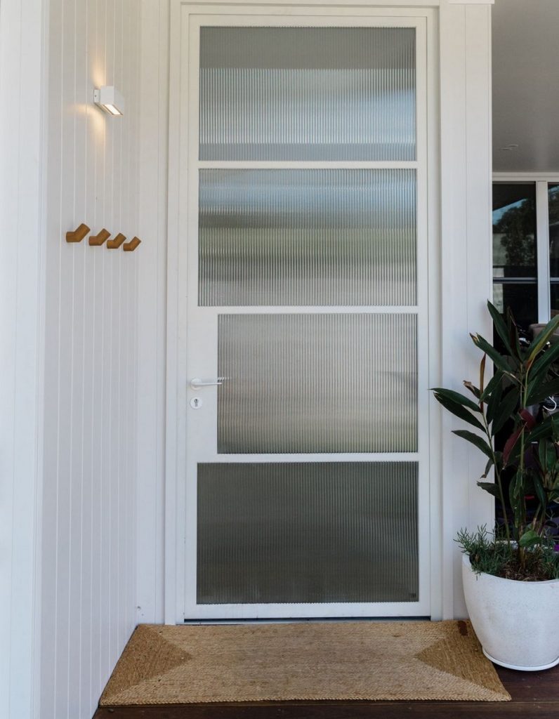 Bespoke steel entry door with frosted glass panels, flanked by a planters and a coir mat, set against a light wooden facade.