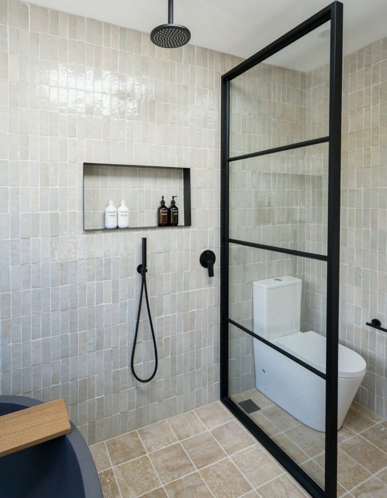 Modern bathroom featuring a sleek black-framed shower enclosure, neutral tiled walls, and a minimalist toilet design, accented by natural light.