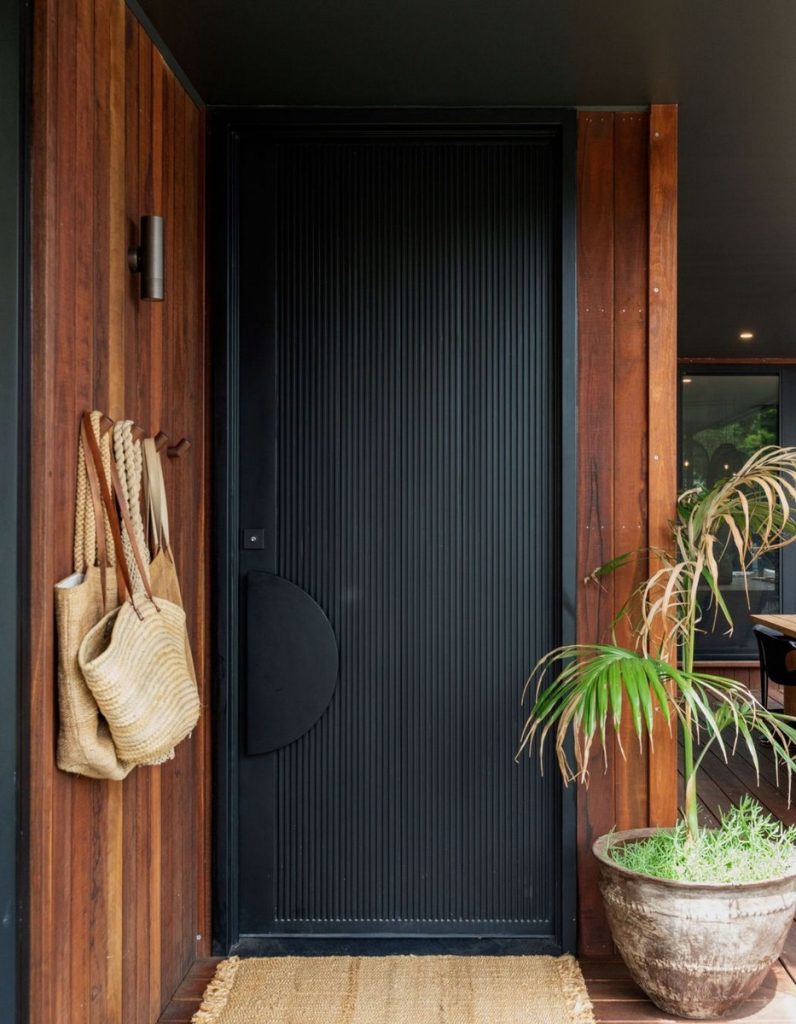 Sleek black ribbed door set against warm timber panelling, complemented by a jute mat, potted plant, and woven bags.