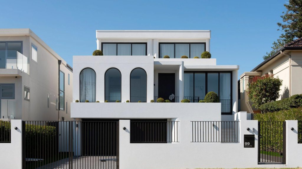 Contemporary two-storey white house with arched windows, minimalist design, and manicured greenery, enclosed by a black fence.