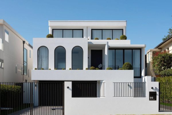 Contemporary two-storey white house with arched windows, minimalist design, and manicured greenery, enclosed by a black fence.