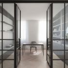 Modern interior featuring sleek steel-framed glass doors, leading to a minimalist workspace with a table and shelves, softly lit by natural light.