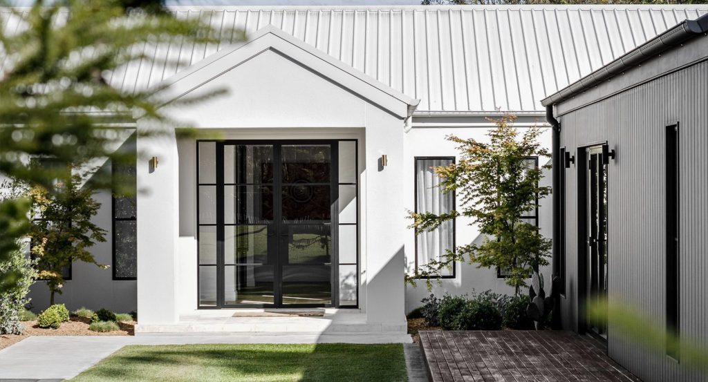 Modern farmhouse entrance featuring a sleek black steel door, white walls, a gabled roof, and neatly landscaped greenery.