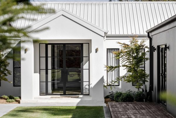 Modern farmhouse entrance featuring a sleek black steel door, white walls, a gabled roof, and neatly landscaped greenery.