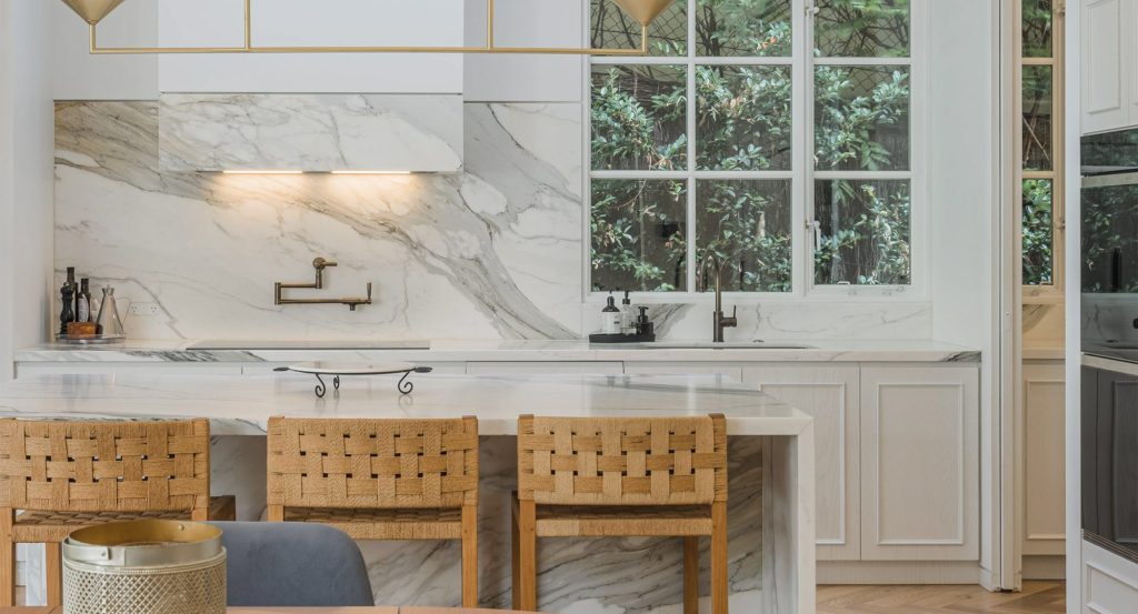 Modern kitchen featuring a marble backsplash, wooden bar stools, and sleek brass fixtures, surrounded by large windows with greenery outside.
