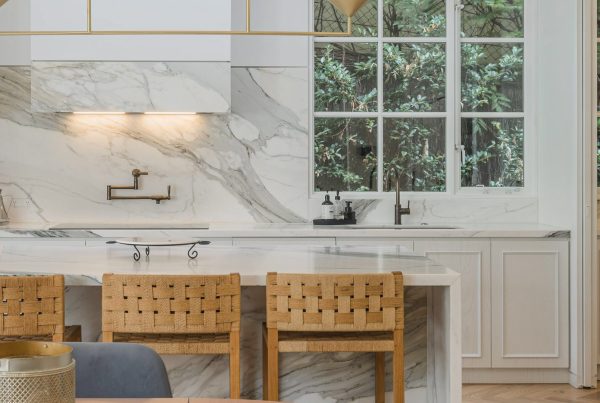 Modern kitchen featuring a marble backsplash, wooden bar stools, and sleek brass fixtures, surrounded by large windows with greenery outside.