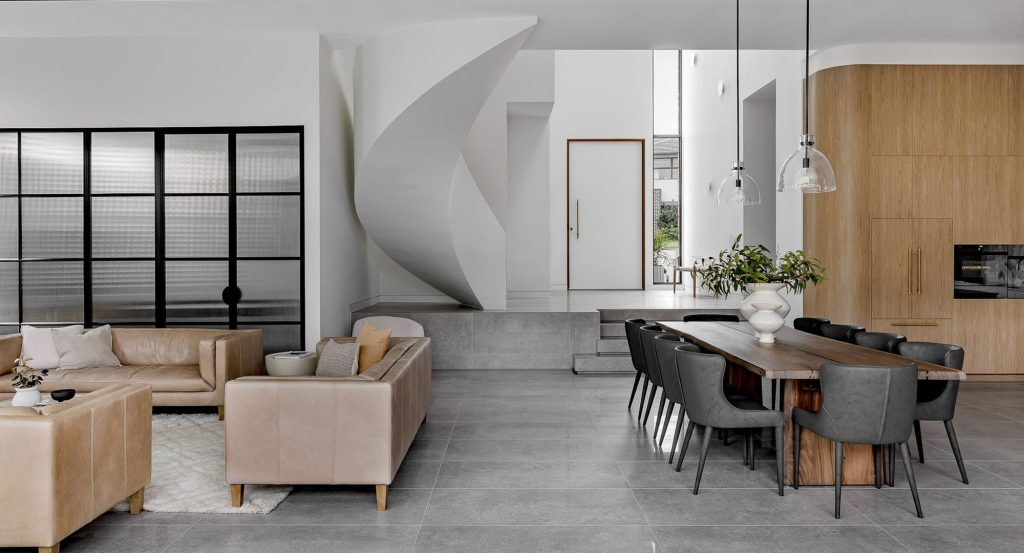 Modern open-plan living area featuring a spiral staircase, stylish beige sofas, a wood dining table, and large steel-framed windows.