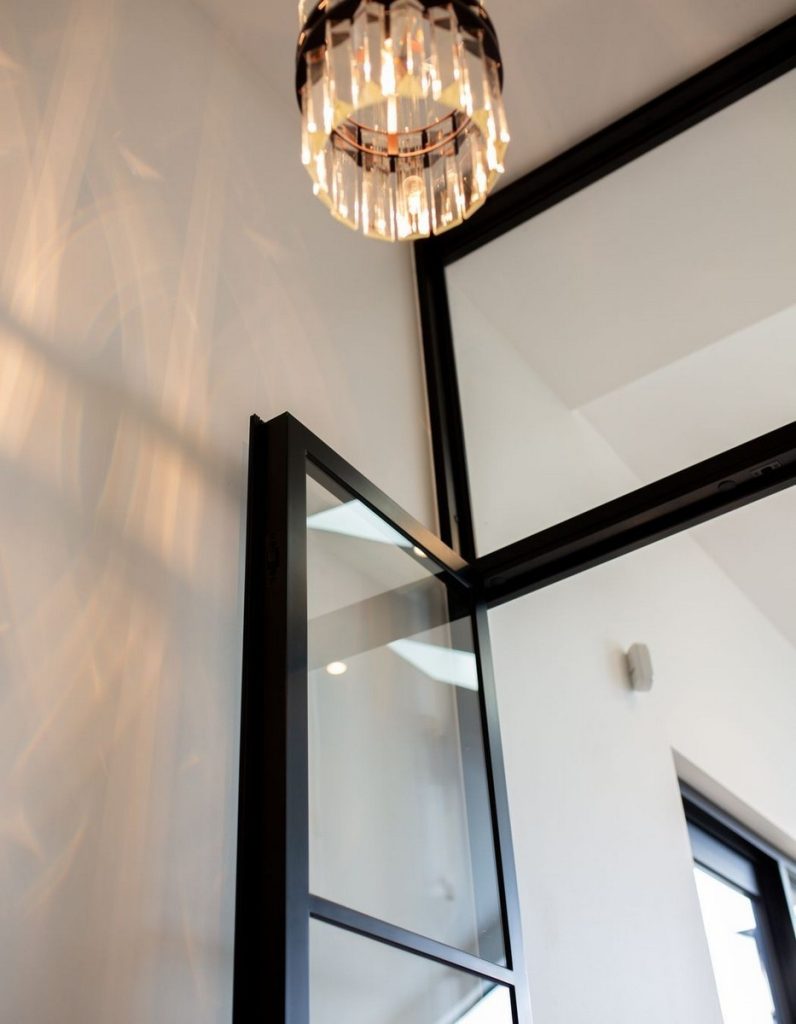 Elegant black steel door with glass panels, illuminated by a decorative crystal chandelier, set against a light-textured wall.