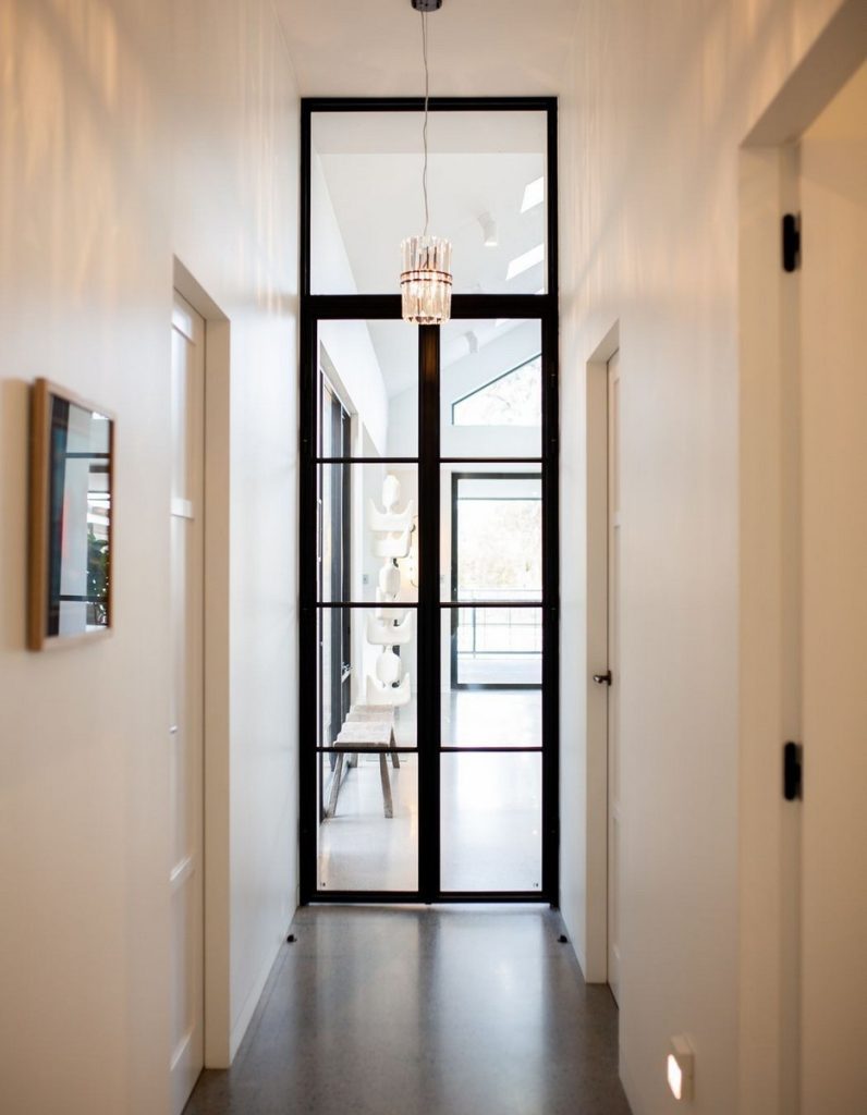 Bright corridor featuring sleek black steel-framed glass doors, white walls, and a modern chandelier above, leading to an airy space.