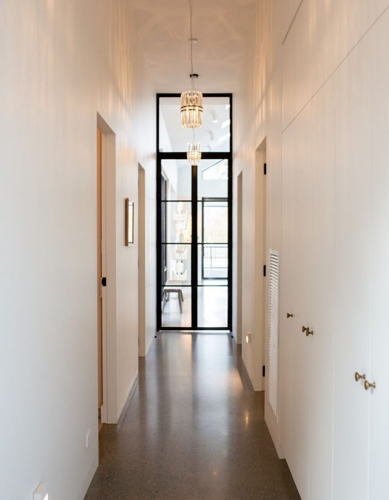 Bright hallway featuring a sleek design with white walls, polished concrete floor, and black-framed glass doors, illuminated by modern pendant lighting.