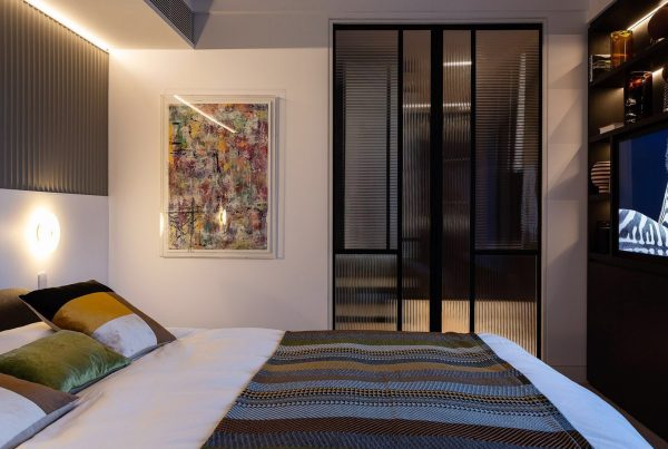 Contemporary bedroom featuring a patterned bedspread, decorative cushions, an abstract artwork, and a stylish glass partition.