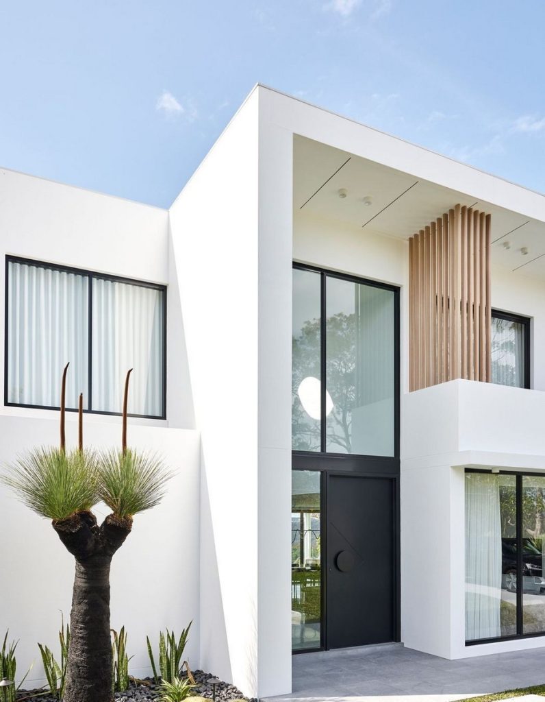 Contemporary white residence featuring large steel windows, wooden accents, and lush greenery, set against a clear blue sky.