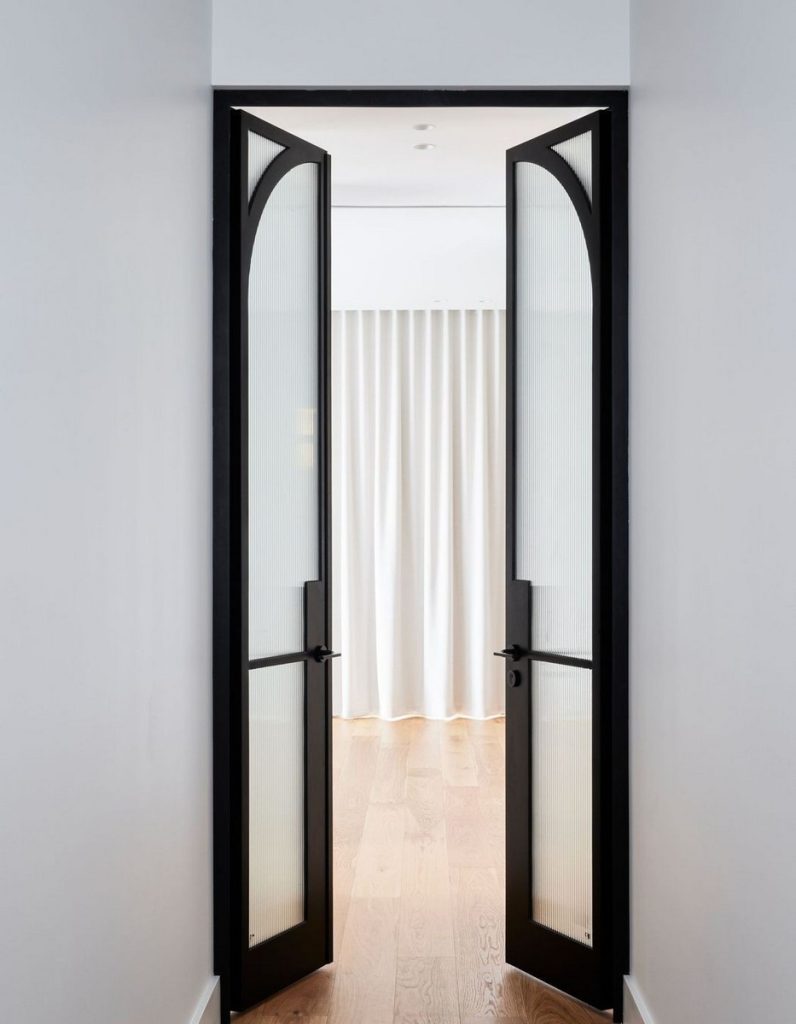 Elegant double doors with a black steel frame lead to a soft-lit space with sheer curtains, showcased against a light wooden floor.