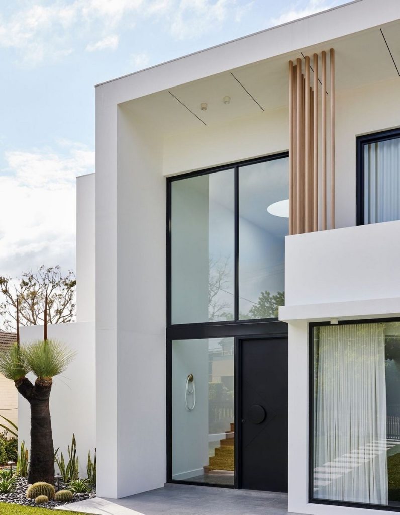 Modern white residence featuring large black steel-framed doors, natural wood accents, and landscaped greenery, displaying architectural elegance.