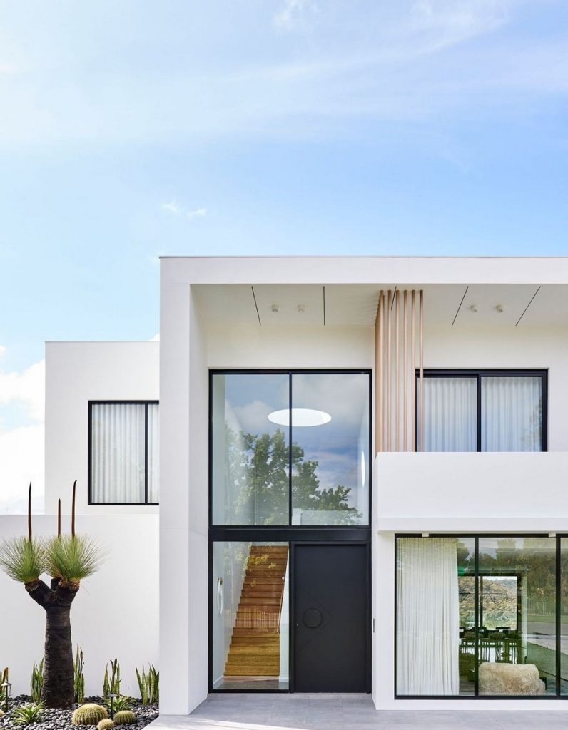 Modern white residence featuring large steel-framed windows, a sleek black door, and green landscaping against a clear blue sky.