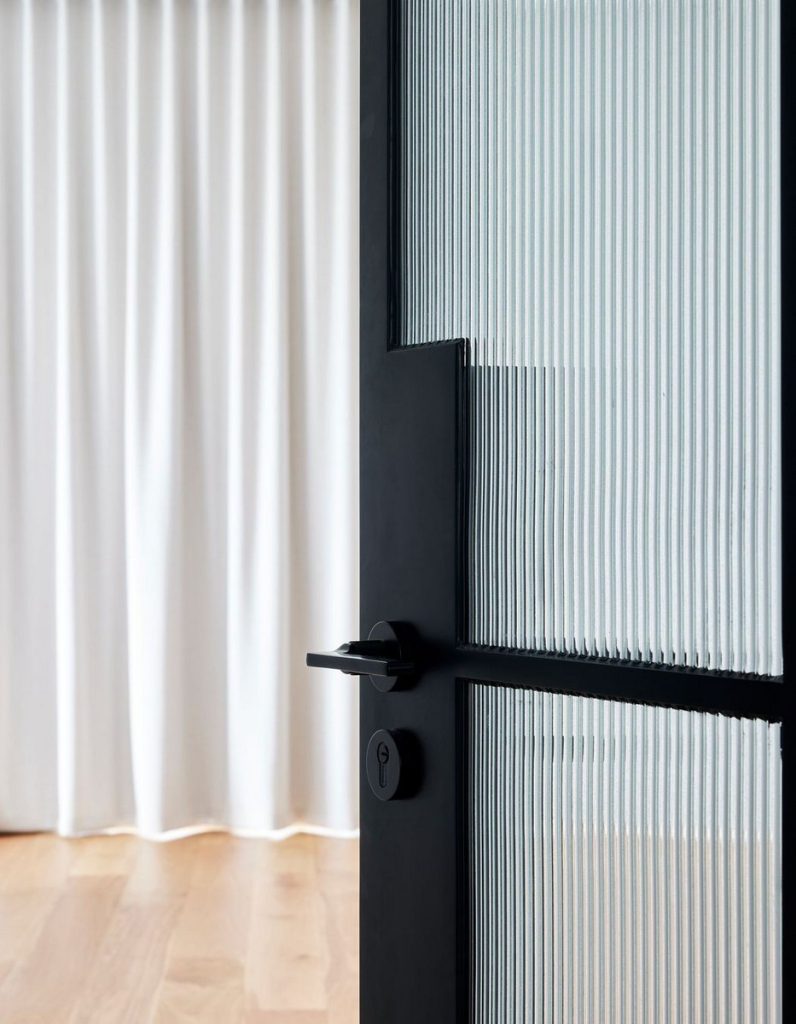 Black steel door with ribbed glass panel open to a softly lit interior featuring sheer white curtains and wooden flooring.