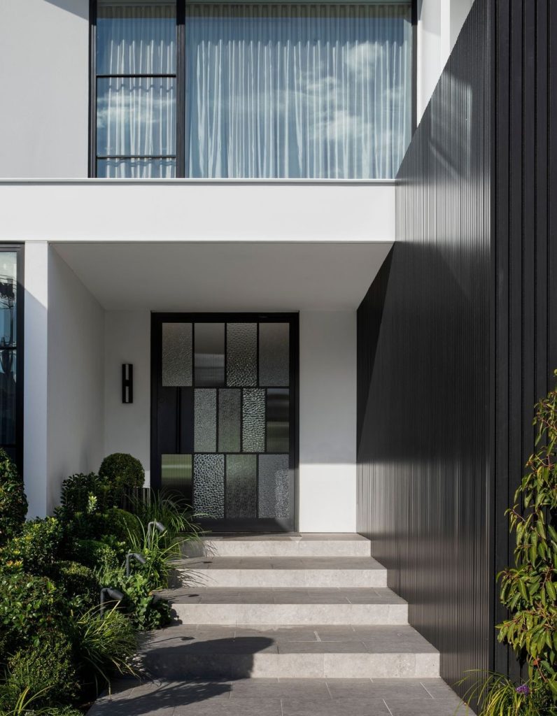 Modern entrance of a stylish white residence featuring a black steel door, large glass panels, and elegant landscaping with steps.