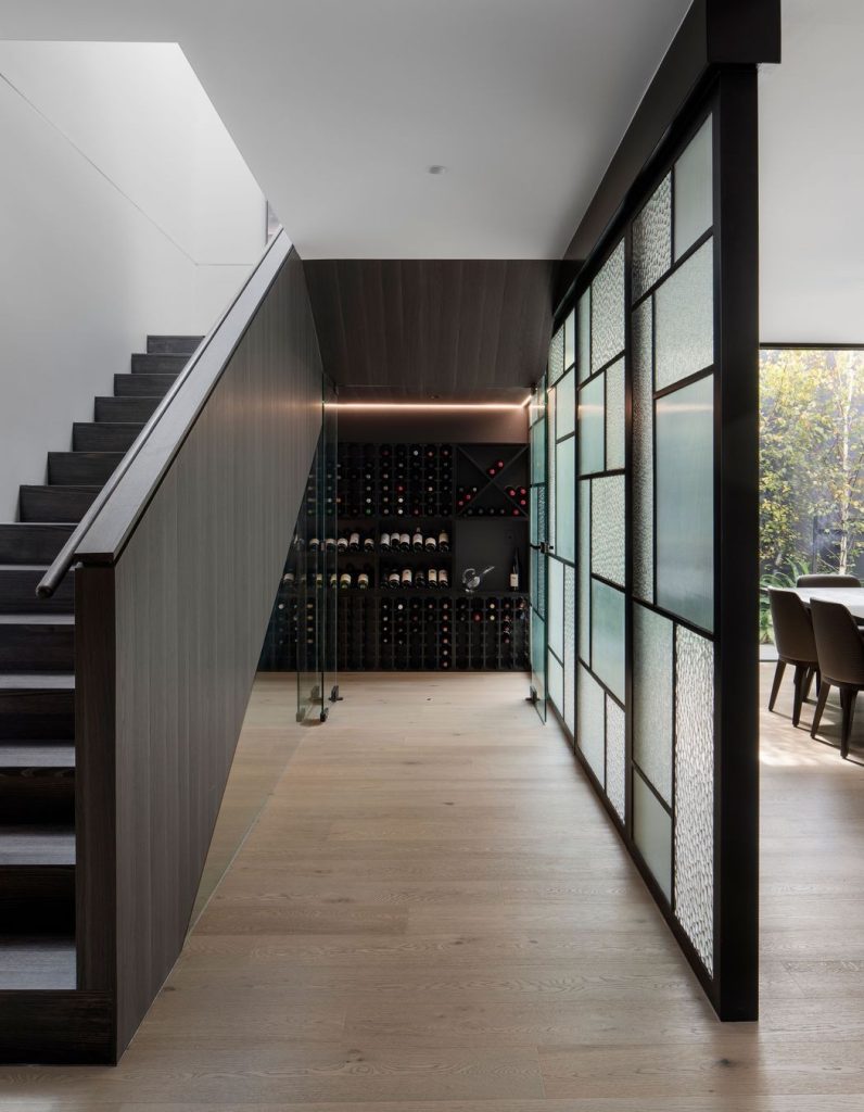 Modern interior featuring a staircase, wine storage wall, and a glass-panelled door, complemented by natural light and wooden flooring.