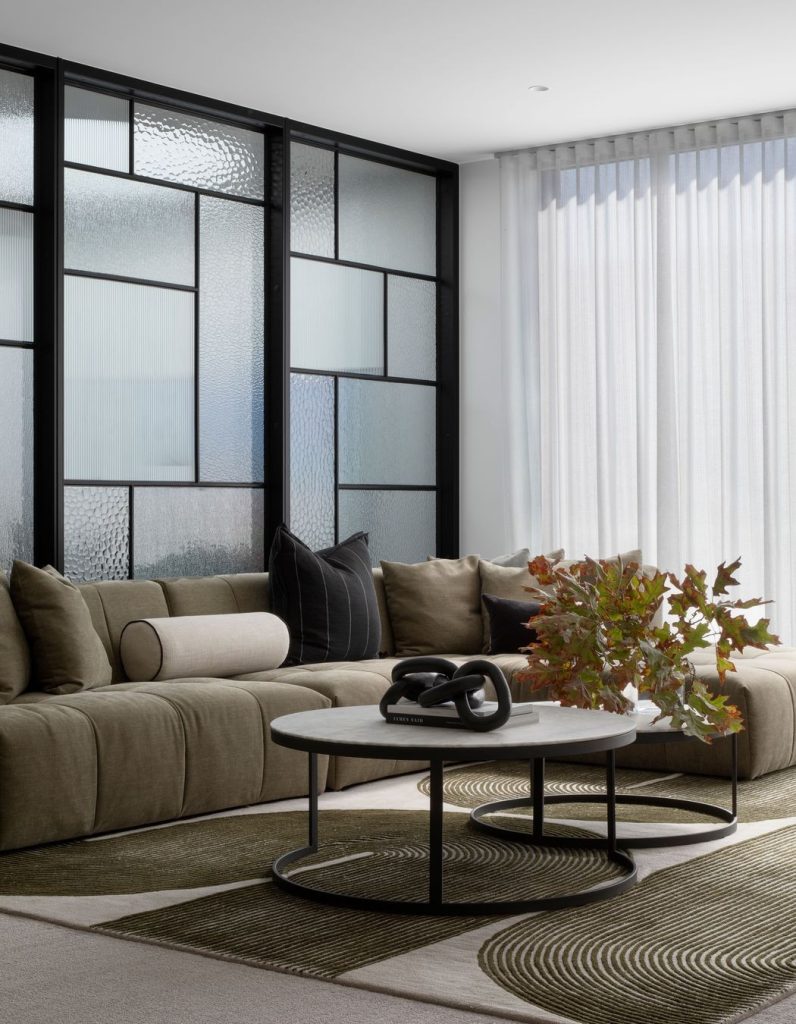 Modern living space featuring a plush olive green sofa, a round marble coffee table, and geometric glass partitions, illuminated by soft drapery.
