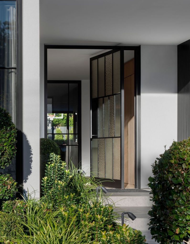 Black steel-framed door with textured glass, opening onto a modern entryway surrounded by lush greenery and neatly trimmed hedges.