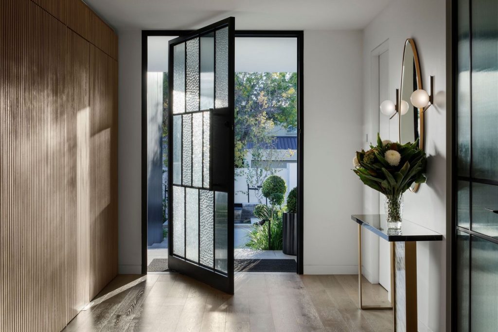 Bright, modern entrance with a large steel-framed glass door, wooden panelling, and a stylish console table adorned with flowers.
