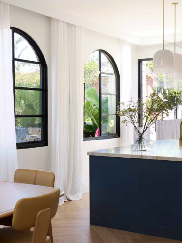 Stylish kitchen featuring arched black steel windows with sheer curtains, a marble countertop, and a wooden dining table with yellow chairs.