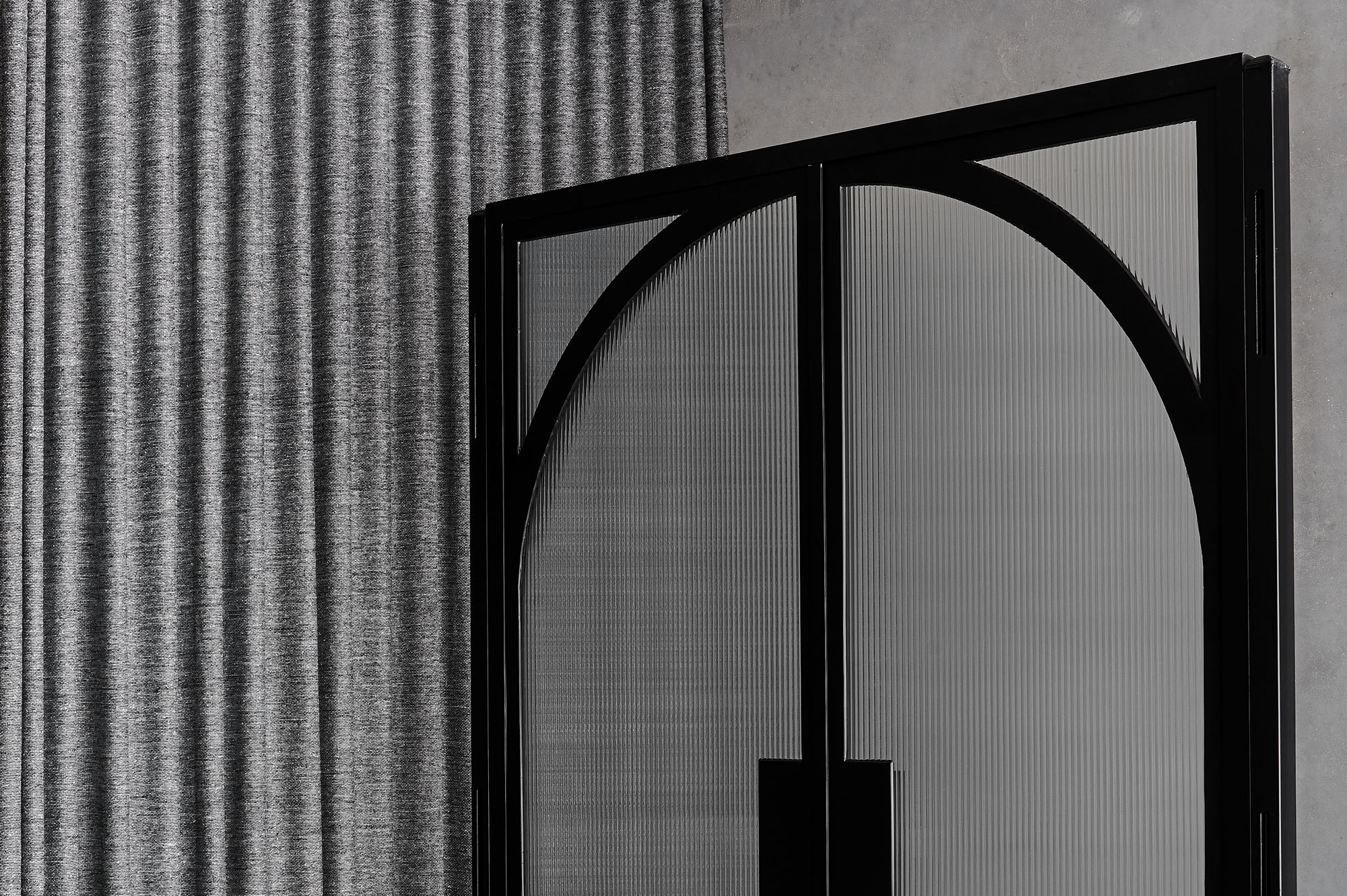 Black steel-framed glass hinge doors with an arched design, set against a textured grey wall and vertical striped curtain.