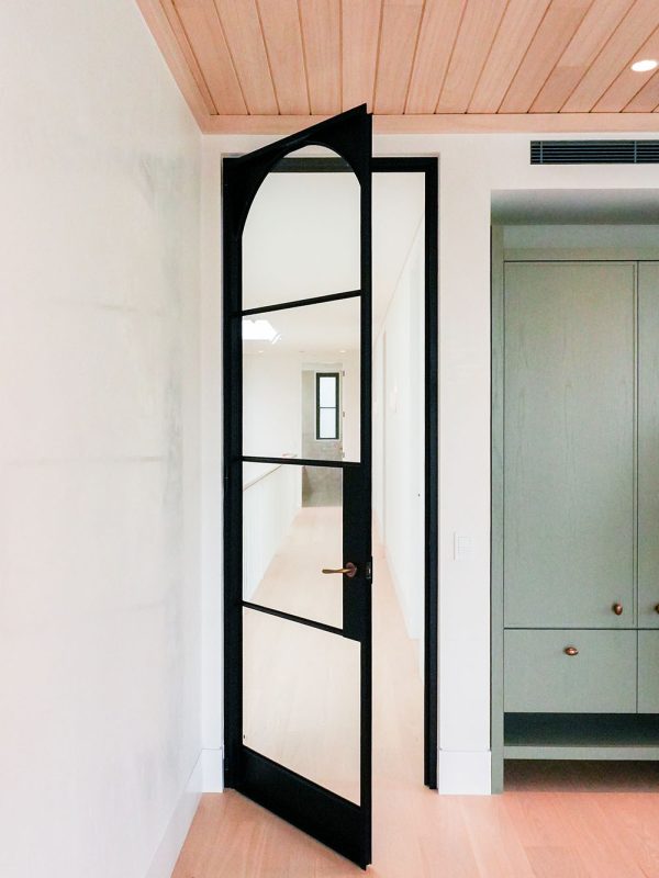 Black steel-framed glass door with an arched top, partially open, leading into a bright interior featuring light wood flooring.