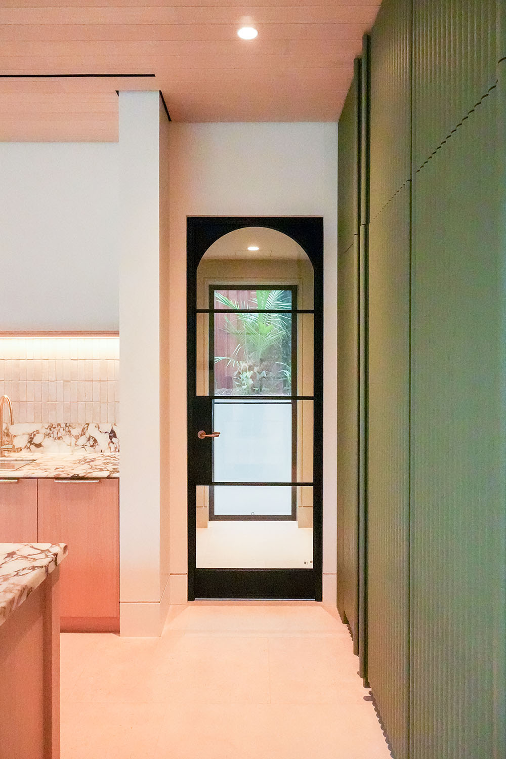 Steel-framed glass hinge door in a modern interior, featuring a black frame, arched top, and surrounded by light-coloured walls and cabinetry.