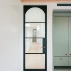 Black steel-frame glass hinge door with an arched top, leading to a light-filled corridor; neutral wood ceiling and green cabinetry visible.