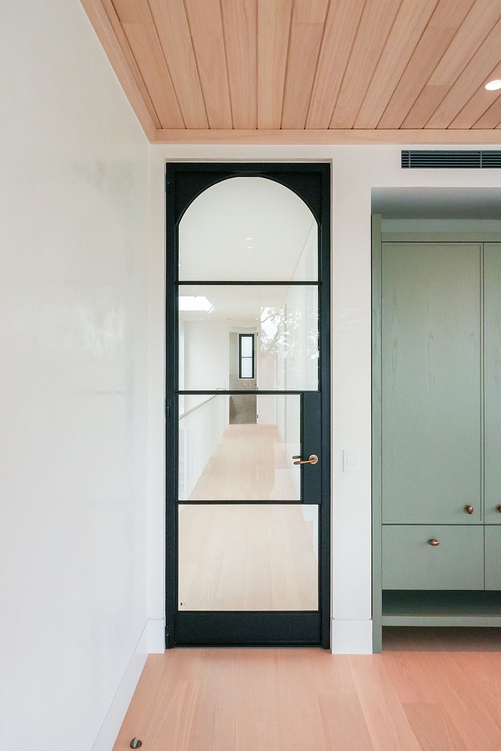 Black steel-frame glass hinge door with an arched top, leading to a light-filled corridor; neutral wood ceiling and green cabinetry visible.